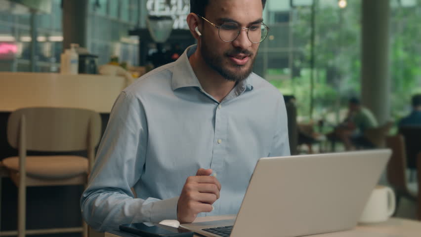 Smiling client tourist male guy Indian Arabian man businessman business worker in earphones chatting video call laptop conference meeting online friendly communicate in cafe computer distant studying