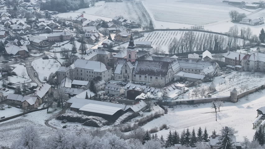 AERIAL: Picturesque small town with famous Sticna monastery covered in snow. Prominent church steeple rises above an old abbey and quaint little village nestled in a snowy winter rural landscape.