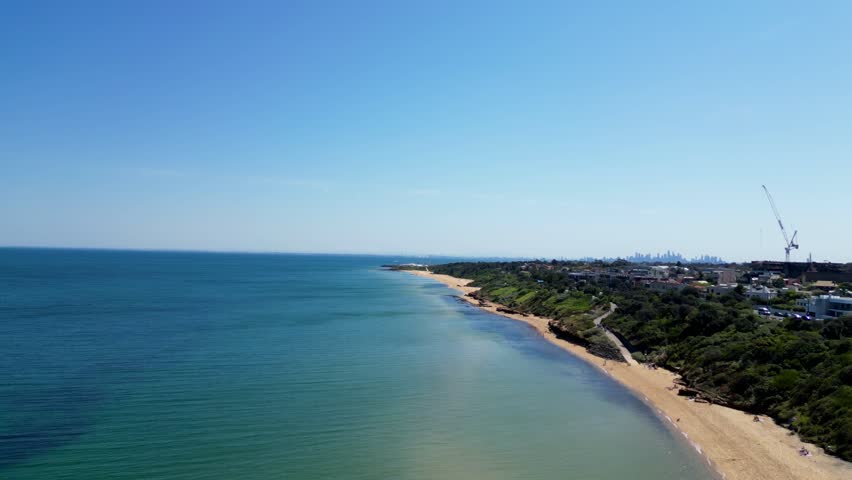 Aerial drone footage of Melbourne beaches with city skyline in the background. Clear view of coastline, sandy shores, and ocean waves meeting urban landscape.