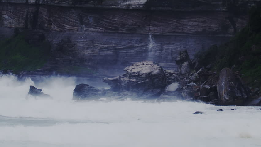 Large waves crashing on a rocky shoreline in Sydney, Australia, with turbulent water and mist.