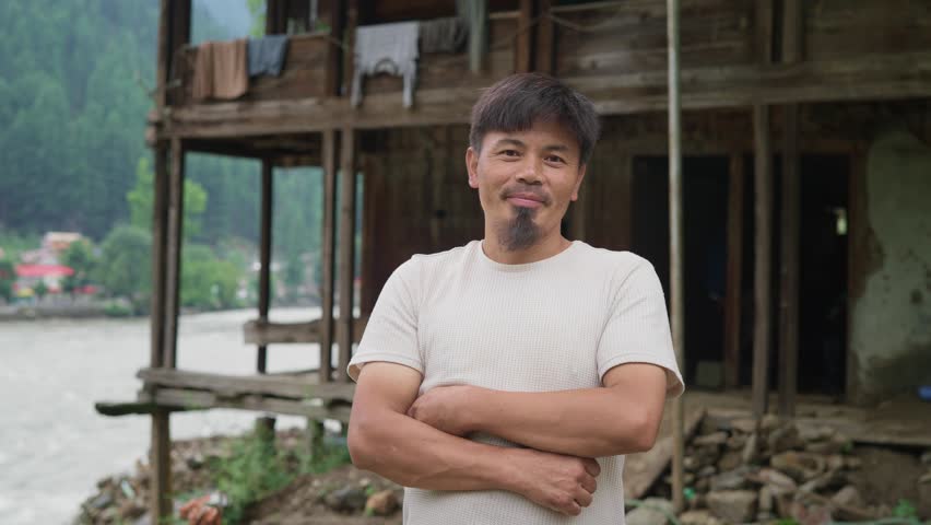 A young happy Asian male or man with crossed arms stands outdoors in a rural area or the countryside in front of a traditional wooden house facing or looking at the camera and smiles 