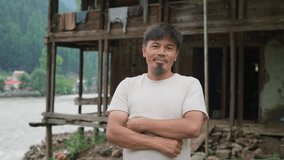 A young happy Asian male or man with crossed arms stands outdoors in a rural area or the countryside in front of a traditional wooden house facing or looking at the camera and smiles  - Powered by Shutterstock - Get 15% off with code: PIKWIZARD15