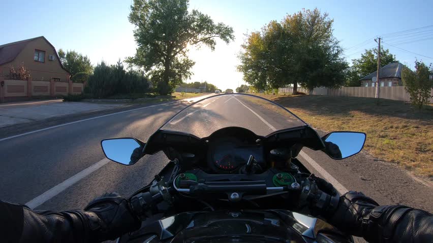 Point of view of a motorcycle rider rides in rural road through village. The viewpoint of motorcyclist driving motorbike at suburban country route. Guy racing his motor bike during summer journey. POV