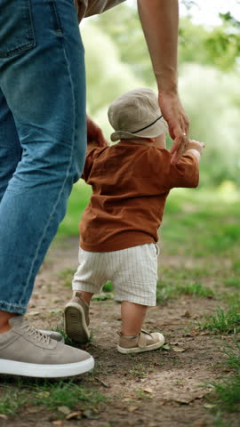 Toddler boy learning to walk supported by his dad. Father holds his son by the hand as the kid steps by the grass. Vertical video.