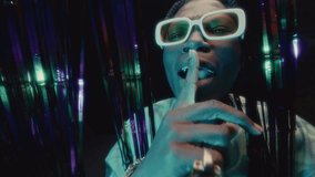 Black hip-hop artist in white-framed glasses and gold accessories rapping to camera behind shimmering tinsel curtain illuminated by colorful lights in nightclub - Powered by Shutterstock - Get 15% off with code: PIKWIZARD15