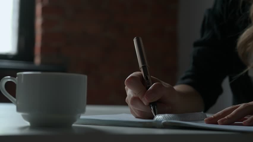 Business woman sitting at office desk writing taking notes in notebook with pen, concentrated person writing down ideas and planning events, close up shot.