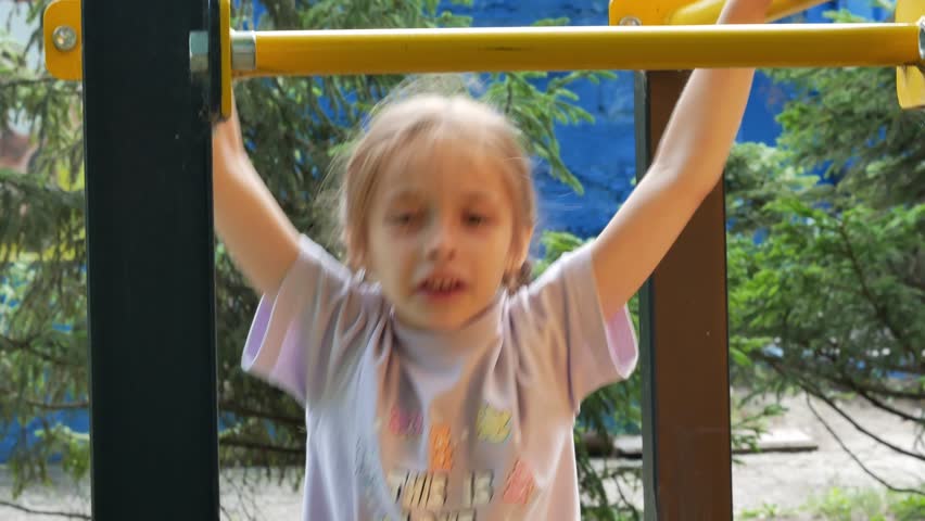 A little girl with pigtails climbs on the playground on the street, threw her legs over the Turks, hanging upside down. A happy childhood. A sporty, active child.