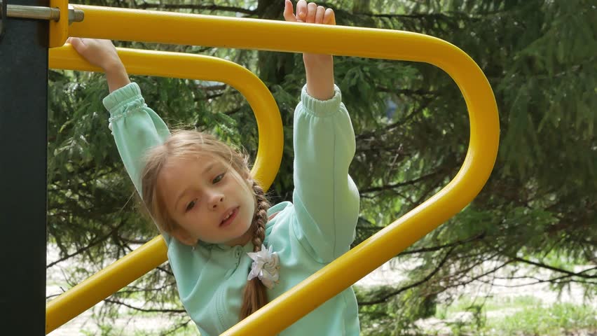 A little girl with pigtails climbs on the playground, pulls herself up and hangs on her hands, overcomes an obstacle, climbs up. A happy childhood. A sporty, active child.