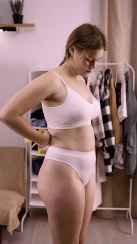 Young woman in underwear measures her breasts with a yellow tape measure at home interior