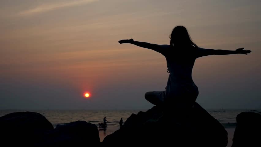 Silhouette serene woman meditating on tranquil sunset ocean beach. Exercise and Healthy lifestyle. A female yogi meditating at beach. Zen Mental health.