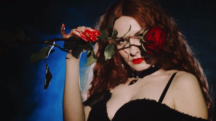 fantasy vampire woman drops of blood fingers hands holding red rose flower, lips evil beauty face pale skin sexy girl white teeth fangs. halloween holiday make-up Gothic lady art night room full smoke