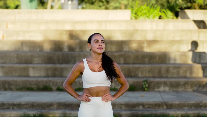 Active woman pausing after workout, hands on hips as catches breath. Determined jogger resting post run, gazing into distance