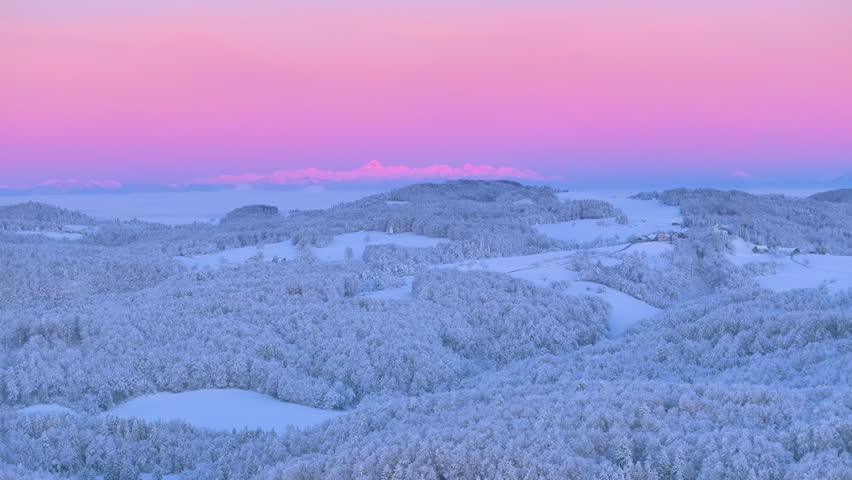 AERIAL: Soft pink sky over snowy forested hills with high Alpine mountains glowing at dawn in the background. Beautiful start to the cold winter day over a hilly landscape freshly whitened by snow.