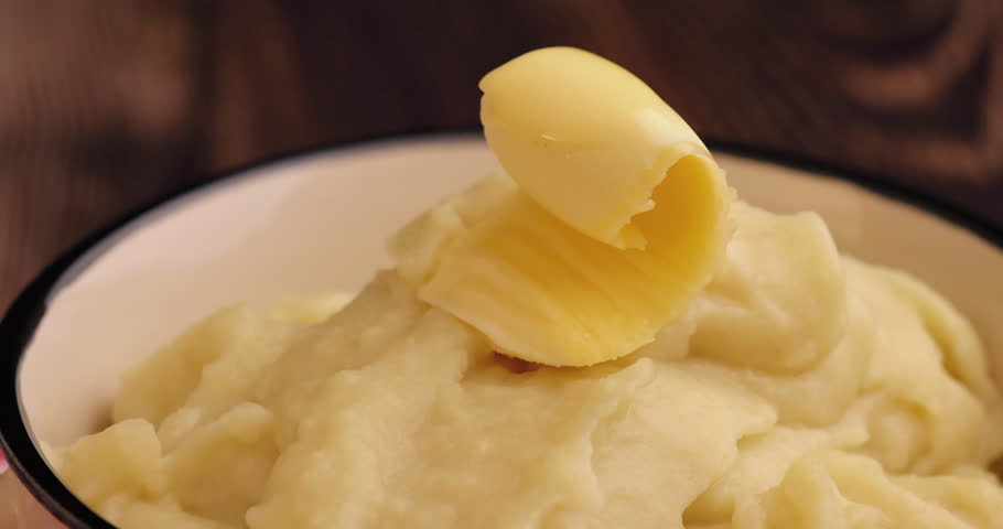 Video. Close-up of mashed potatoes with melting butter slice in a rustic wooden bowl on an old wooden table, evoking warmth and traditional comfort food