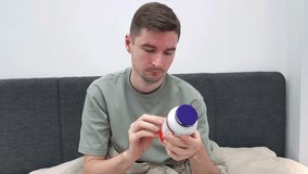 Man reading label on white plastic supplement bottle while sitting in bed. Man holding a supplement capsule and examining it with a bottle in hand - Powered by Shutterstock - Get 15% off with code: PIKWIZARD15