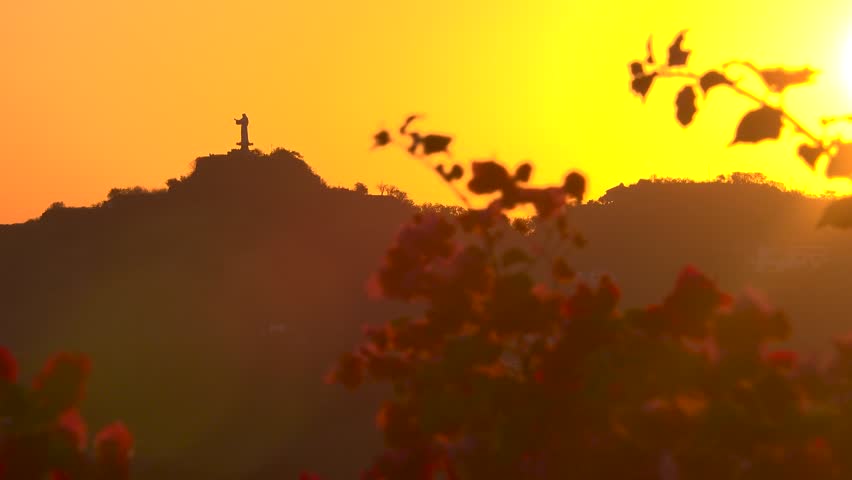 A statue of Jesus on a hill during a beautiful sunset. Red flowers in the foreground at San Juan del Sur, Nicaragua