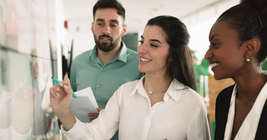 Latina woman team leader explain information presented on white board, provide data to diverse male female teammates, work together on joint presentation, develop marketing strategy, planning start up