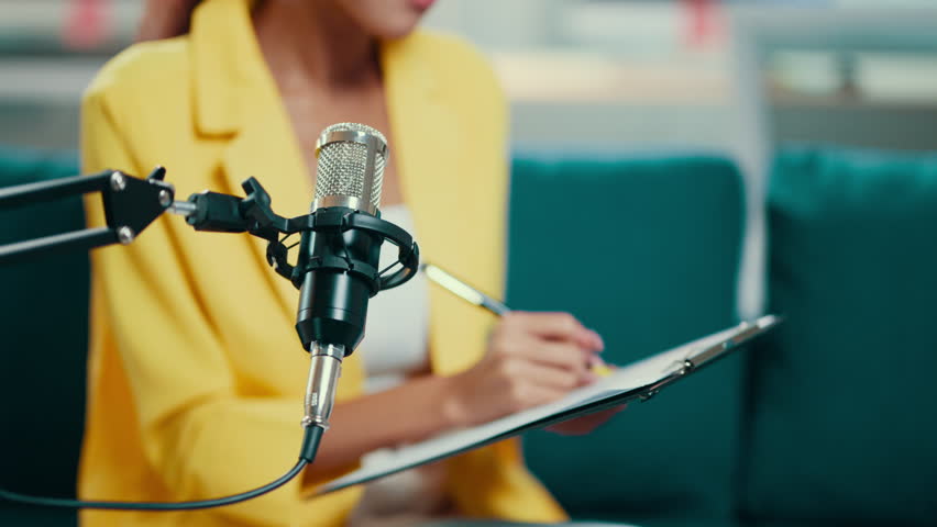 African american radio host wearing a yellow blazer is sitting and taking notes on a clipboard while recording a podcast in a radio station