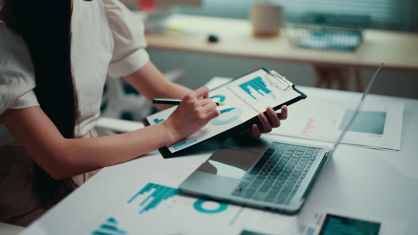 Businesswoman is sitting at her desk and analyzing financial charts on a clipboard. She is using a pen to point at the data