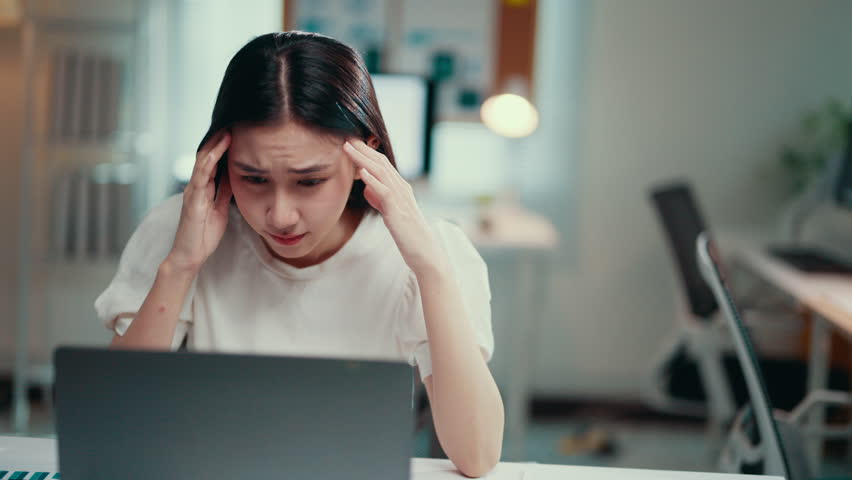 Young asian freelancer woman is massaging her temples, suffering from headache while sitting at the desk and working remotely on a laptop from home