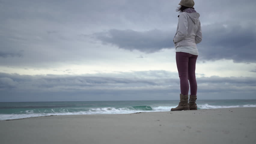A solitary woman on an empty beach gazes at the ocean under a cloudy sky. Waves crash, symbolizing the loneliness and social isolation of modern life. Reflective and melancholic mood. 4K.