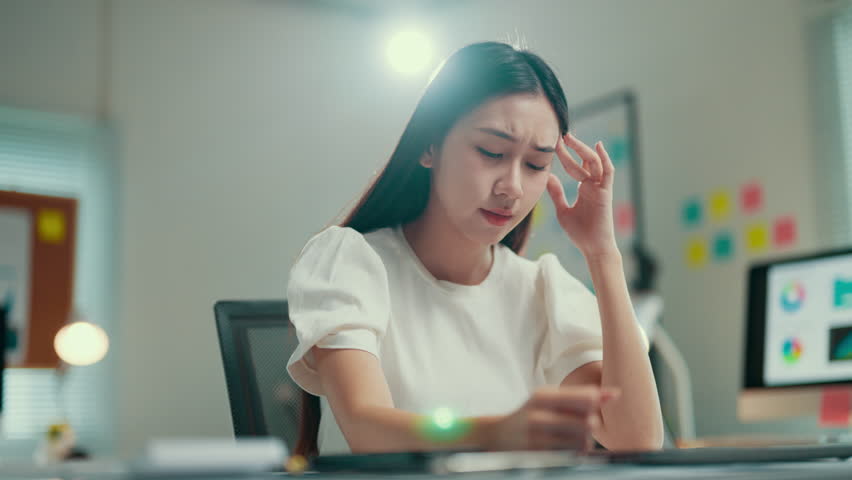 Young asian businesswoman is massaging her temples, trying to relieve a painful headache. She is sitting at her desk in her office