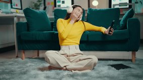 Young woman is sitting on the floor, using her smartphone for a video call. She is wearing headphones and is laughing and smiling - Powered by Shutterstock - Get 15% off with code: PIKWIZARD15