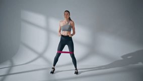 Female athlete doing squats with elastic band on white wall background. Fitness trainer performing exercises for buttocks and legs alone at empty studio. Active woman using resistance rubber indoors - Powered by Shutterstock - Get 15% off with code: PIKWIZARD15