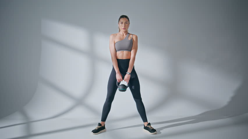 Strong model lifting weight equipment at grey wall background. Young sportswoman bodybuilder making exercise for strength alone indoors. Weightlifting healthy woman enjoying muscle workout in loft gym