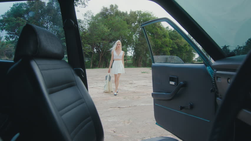 A woman wearing a white dress is strolling towards an open car door in a tranquil rural setting