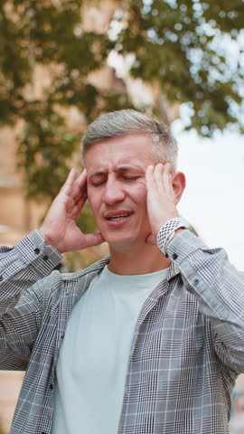 Displeased exhausted Caucasian man rubbing temples to cure headache problem suffering from tension migraine stress. Mature male guy tourist grimacing, pain standing on city urban street. Vertical.