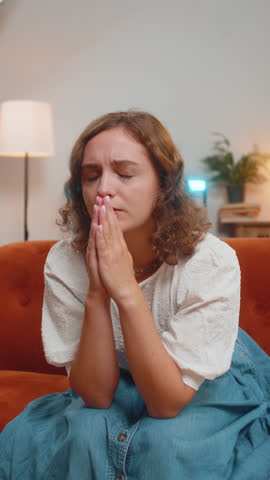 Sad young woman sitting on sofa at home in shock by awful news, folding palms together pray ask for help, thinks feel stressed looks desperate by life problems. Praying, asking for help. Vertical.