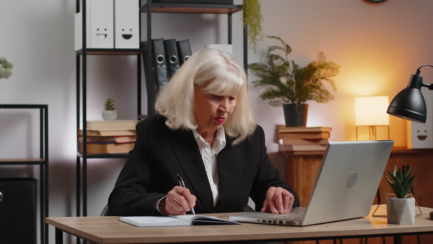 Senior businesswoman writing down or taking down notes while attending online video class or office meeting on laptop at home office desk. Girl freelancer grandma support services looking at webcam. - Powered by Shutterstock - Get 15% off with code: PIKWIZARD15
