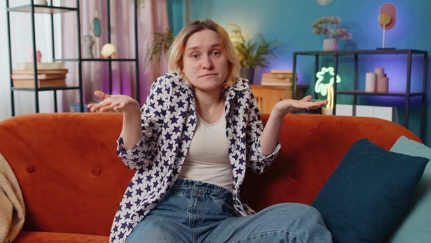 I don't know what to say. Confused Caucasian woman feeling embarrassed about ambiguous question, having doubts, no idea, being clueless and uncertain. Young girl sitting on sofa in living room at home