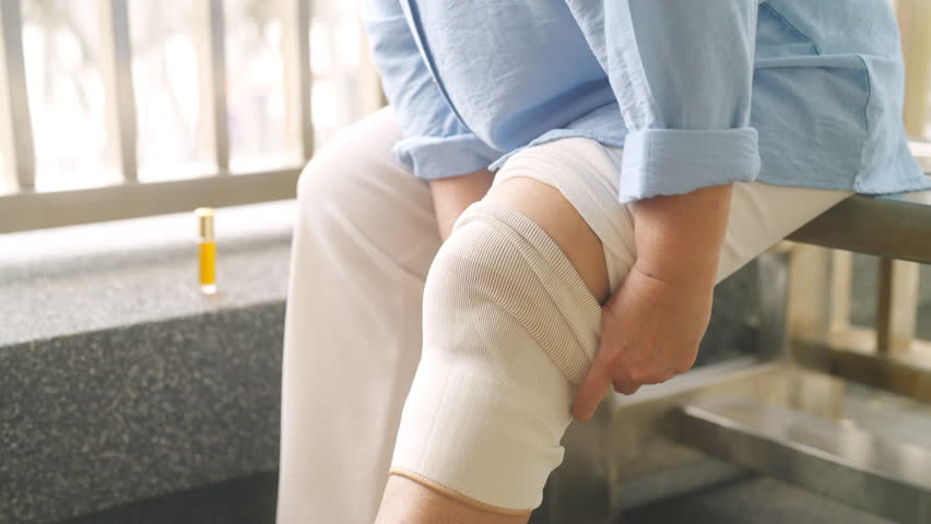 Asian woman get injury knee pain and wearing elastic knee support on leg during travel city. Elderly retired woman travel Thailand on summer holiday vacation. Travel and health insurance concept.