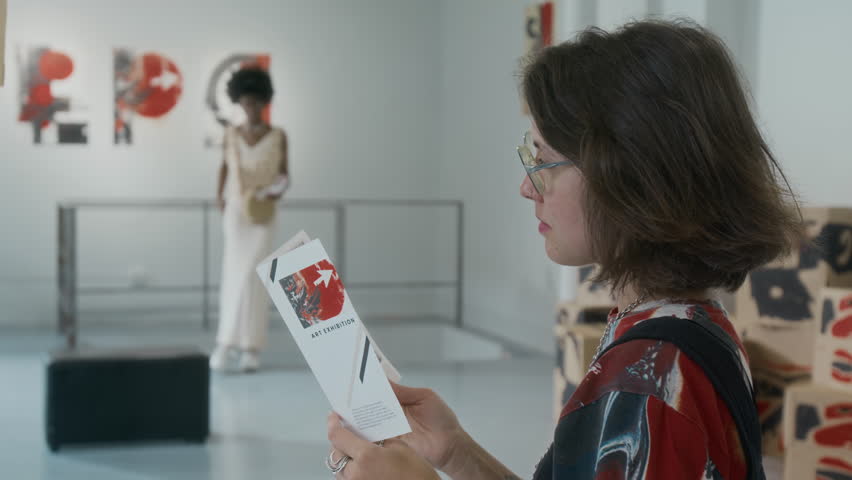 Medium closeup side shot of stylish young Caucasian woman standing in museum at modern art exhibition, viewing abstract paintings and installations, reading brochure, looking around and smiling
