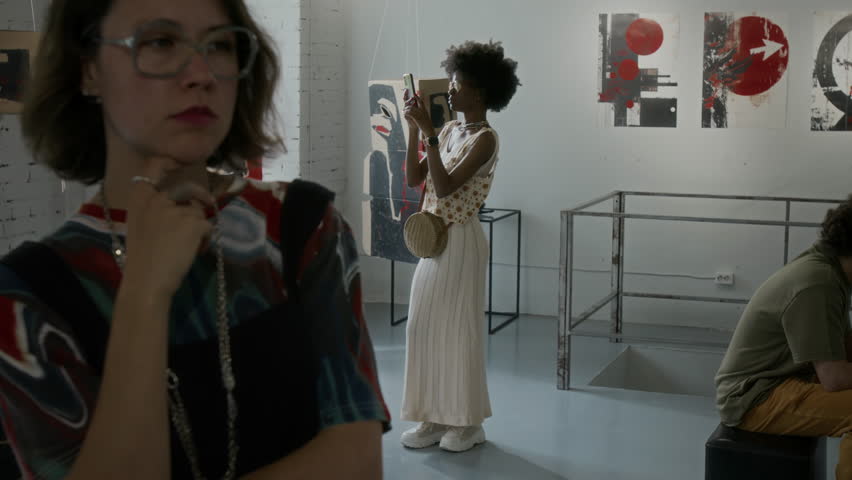 Medium full shot of multiethnic visitors attending contemporary art exhibition in museum - Caucasian woman admiring painting, African American girl taking selfies with artworks, man resting on bench