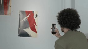 Medium rear shot of young man with thick short curly hair standing in front of artworks displayed on white wall at modern art exhibition and taking photos on smartphone - Powered by Shutterstock - Get 15% off with code: PIKWIZARD15