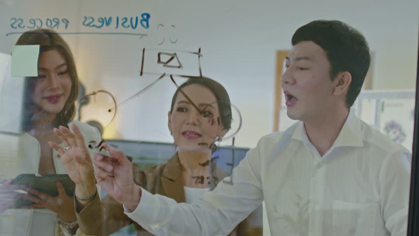 Diverse group of professionals collaborates on project, brainstorming ideas on transparent board in office setting. Showcases teamwork, creative problem-solving, and modern business practices