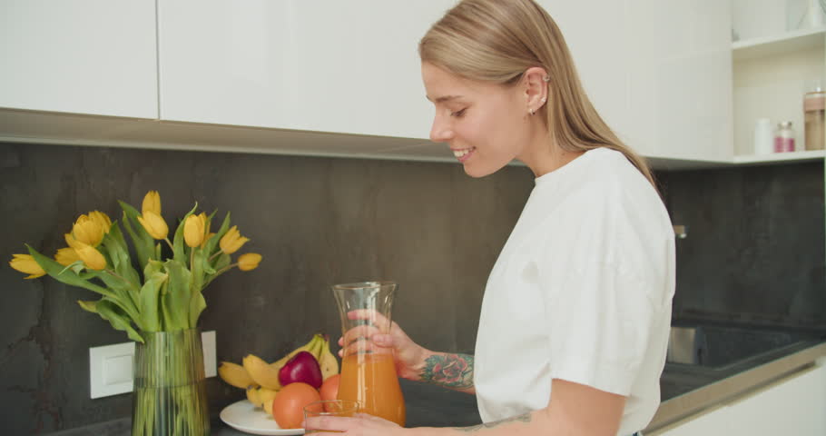 A young woman enjoying a glass of fresh orange juice in a bright modern kitchen, surrounded by vibrant fruits and a peaceful atmosphere