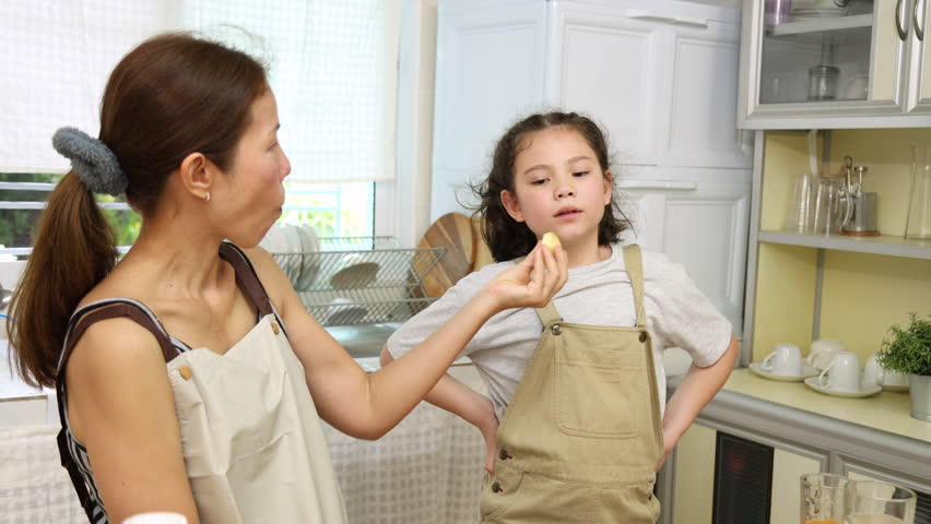 Asian mother and daughter sharing meal in kitchen. young girl enjoys croissant smiling, woman watching kid eating in warmly moment. preparation lunch meal cooking together, family lifestyle at home
