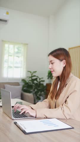 A young woman is sitting at her desk in a modern office, focused on her laptop. She is working in a clean and organized environment, with notebooks and office supplies around her.