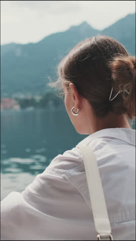 Vertical video, Young brown-haired woman dressed in white shirt enjoying beautiful view of lake and mountains view from behind