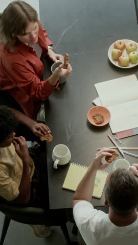 Vertical top shot of multiethnic foster family - Caucasian mom, dad and African American son sitting together at table, enjoying homemade biscuits with tea and chatting