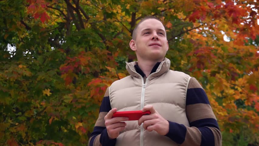 one male person in casual clothes with a cell phone walks on a blurred autumn background and talks via mobile communication.
