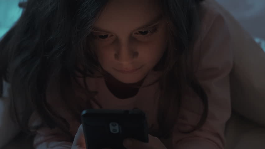 Social media. Gadget use. Child insomnia. Late talk. Internet meeting. Tired bored small girl using mobile phone texting message in bed in dark home bedroom with blue light.
