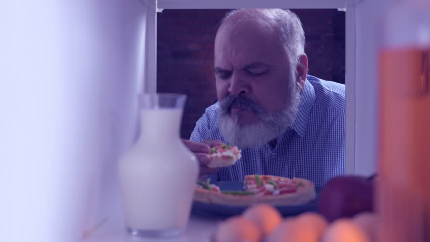 Indian Asian senior businessman with a white beard quietly opening the fridge in the middle of the night, eating a slice of pizza secretely