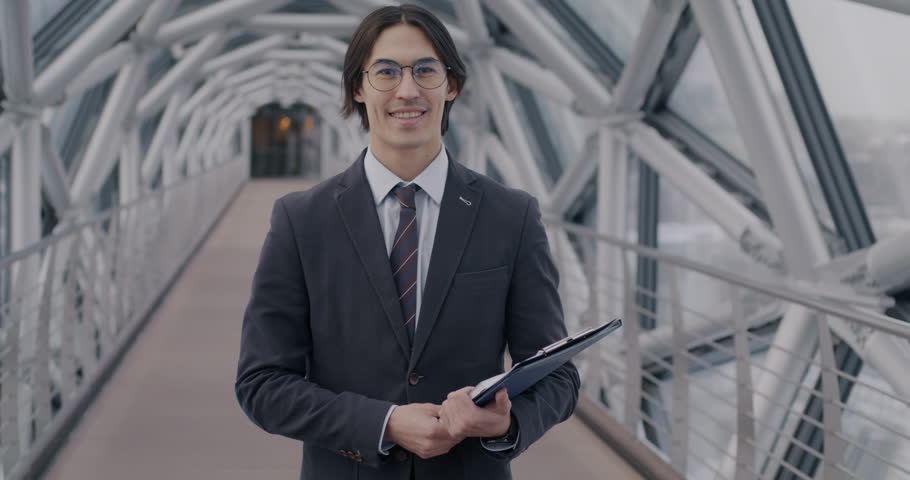 Slow motion portrait of young Asian businessman in suit standing in modern glass hall holding folder with documents smiling looking at camera. Business and people concept.