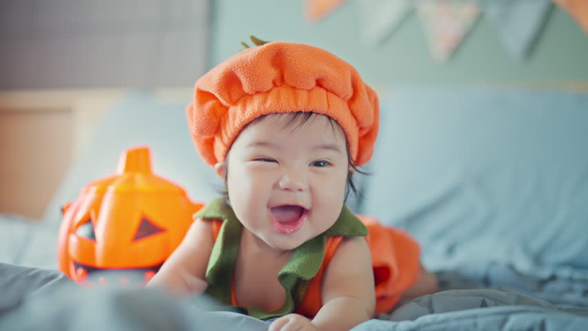 Happy cute asian newborn baby girl dressed in Halloween jack o lantern costume during tummy time on bed with Halloween decoration, having fun and celebrating Halloween at home