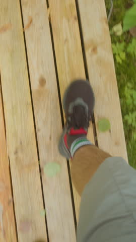 Vertical POV shot of anonymous hiker walking on wood plank trail during hiking journey in lush forest nature at daytime